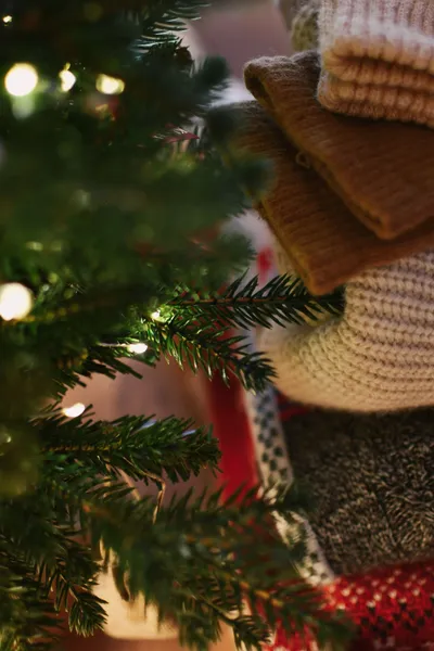 Pile de pulls en maille pliés, beige, camel, écru, gris chiné et jacquard rouge et blanc, à côté d'une branche de sapin éclairée par une guirlande.