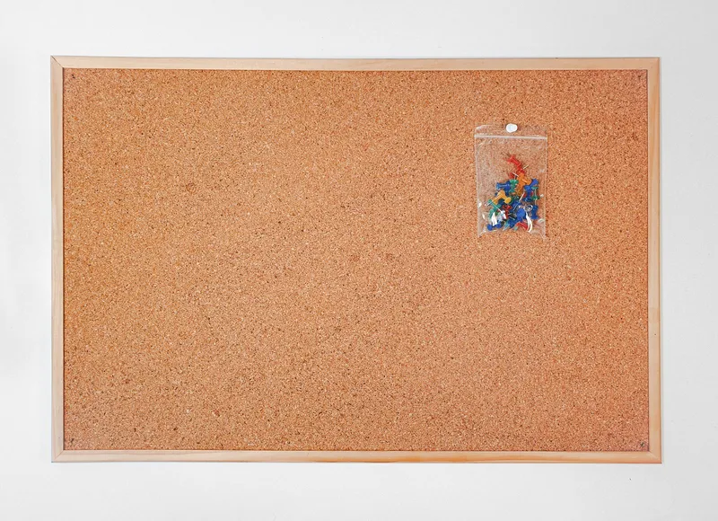 Tableau en liège vierge à cadre en bois clair, avec un sachet de punaises de couleur épinglé dans un coin.