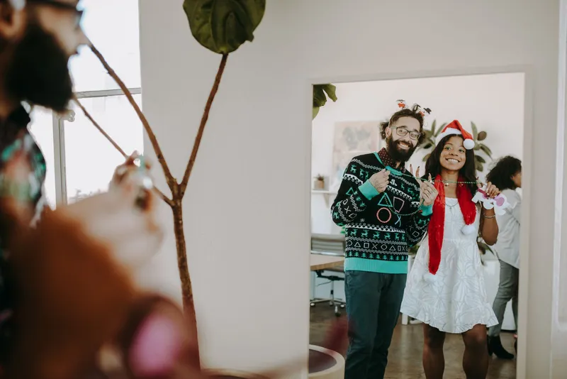 Dans l'encadrement d'une porte de bureau, un homme en pull de Noël jacquard vert et noir, serre-tête de fête, et une collègue au bonnet de Père Noël sourient.