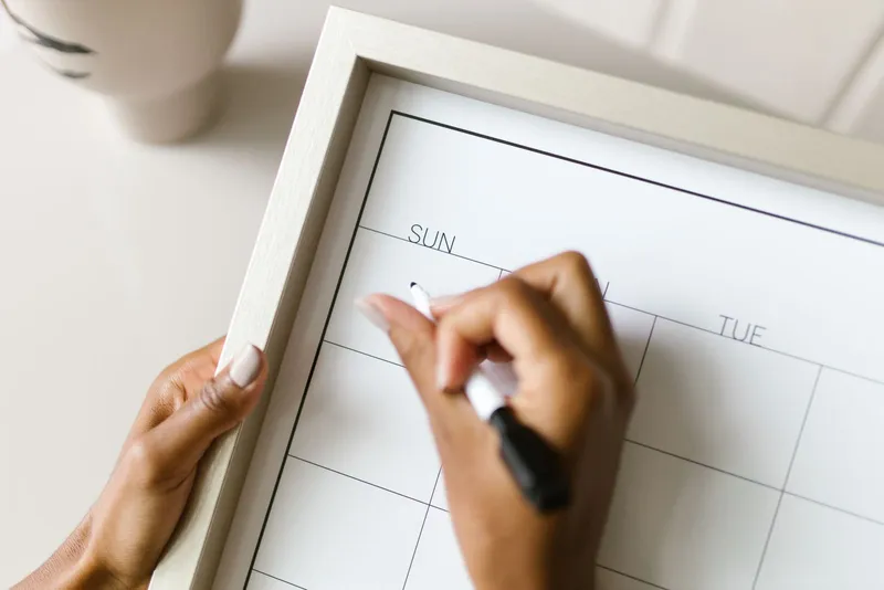 Deux mains inscrivent une date au feutre noir sur un calendrier mensuel vierge, dans un cadre en bois clair.