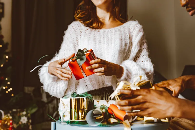 Échange de cadeaux autour d'une table : une personne en pull de laine blanc tient un paquet rouge noué de vert, une autre main défait un paquet doré.