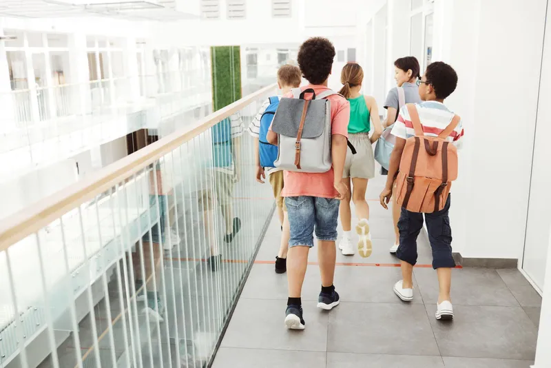 Groupe de collégiens vus de dos, sac à dos sur l'épaule, marchant dans une coursive vitrée et lumineuse de leur établissement.