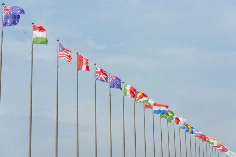 Rangée de mâts portant les drapeaux de nombreux pays, dont le Royaume-Uni, l'Australie et le Brésil, sous un ciel bleu voilé.