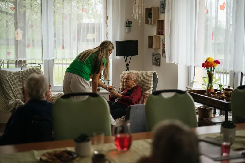 Salle commune d'une maison de retraite : une soignante se penche vers une résidente assise dans un fauteuil, d'autres résidents attablés au premier plan.