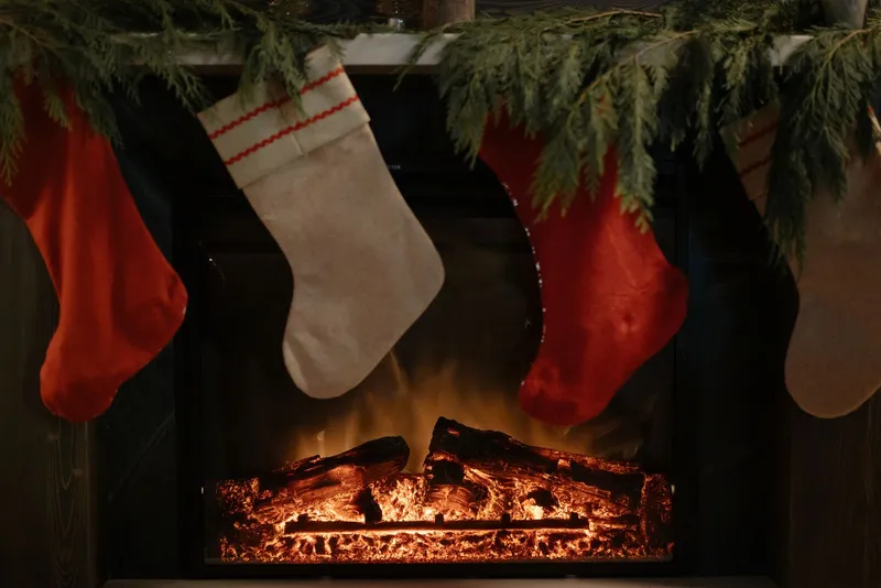 Chaussettes de Noël rouges et en toile écrue suspendues à une cheminée garnie de branches de sapin, devant un feu de bûches.