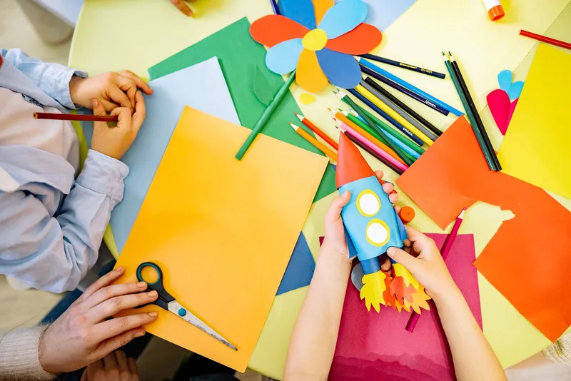 Vue du dessus d'une table de bricolage : mains d'enfants, feuilles de couleur, crayons, ciseaux et une fusée fabriquée avec un rouleau de carton.