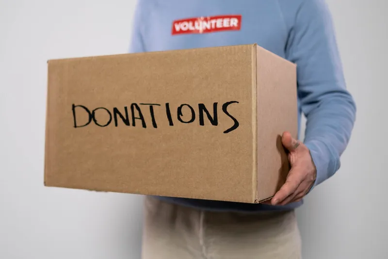 Bénévole en sweat bleu marqué VOLUNTEER portant un carton sur lequel est écrit DONATIONS au feutre noir.