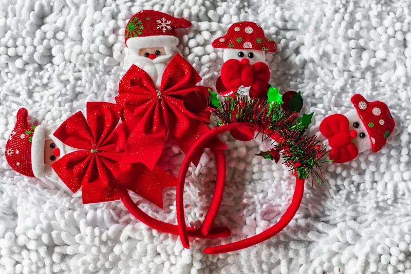 Serre-têtes de Noël rouges ornés de nœuds, de guirlande scintillante, de petits pères Noël et de bonshommes de neige, posés sur un tissu blanc duveteux.